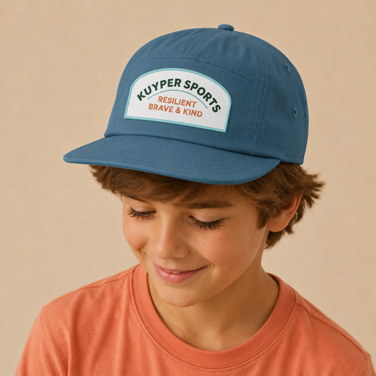A young boy wearing a slate blue baseball cap with a white patch that reads 'KUYPER SPORTS RESILIENT BRAVE & KIND'.